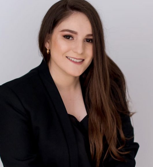 Luana Machado, fonoaudióloga, sorrindo, de cabelo longo castanho e blazer preto.